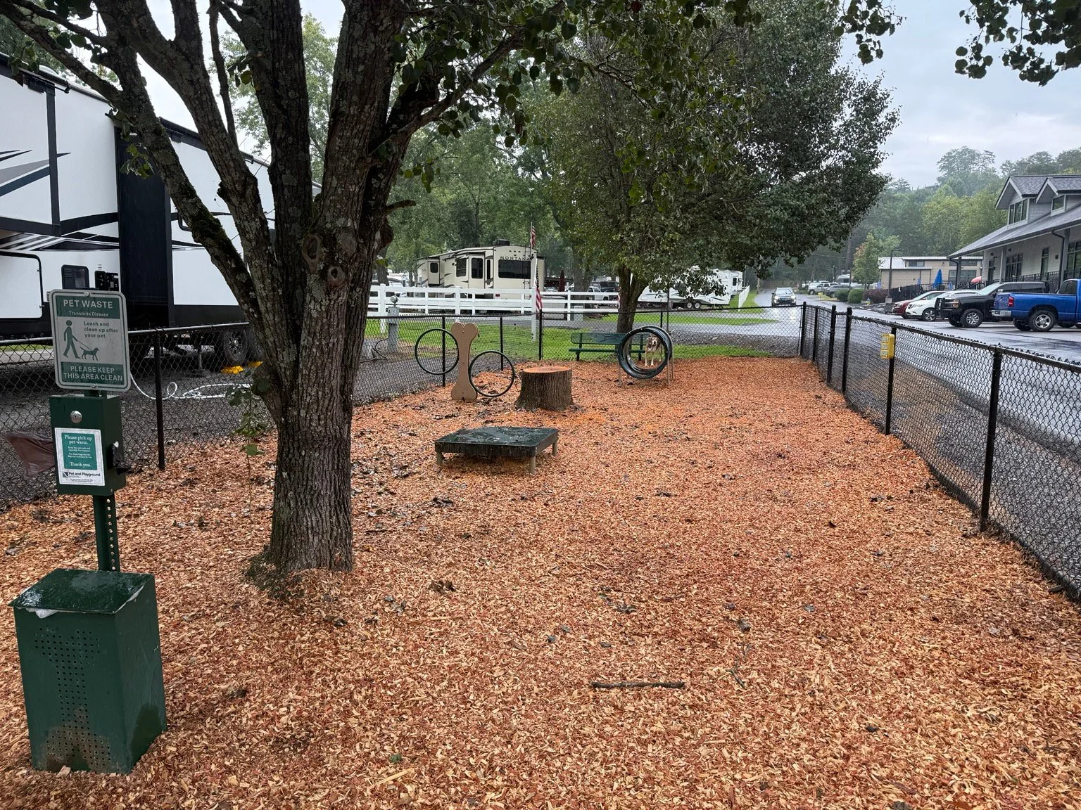Fenced dog park at Clabough's Campground with shade trees, wood chips, hoops, a platform, and a pet-waste station.